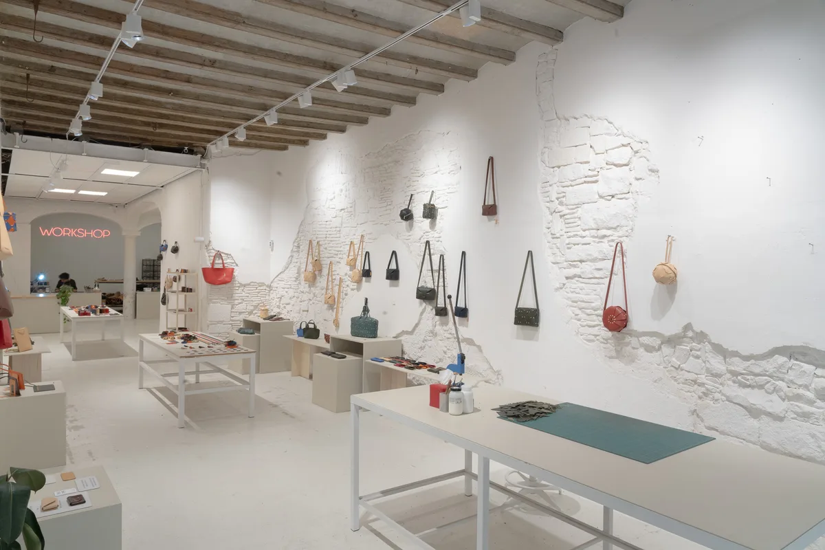 Interior de la tienda de Atelier Madre con bolsos colgados del muro encalado y el rótulo de neón WORKSHOP al fondo de la sala
