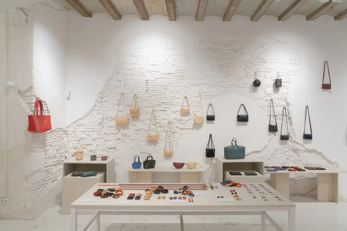 Mesa central de Atelier Madre con pendientes, pulseras y tarjeteros de piel, y bolsos colgados del muro de piedra al fondo